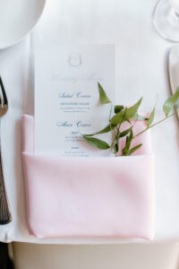 A light pink napkin folded to hold a wedding reception menu and decorative greenery.