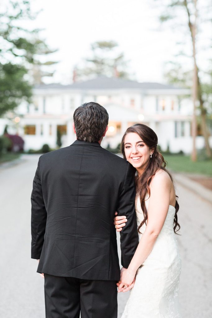 Bride smiling over her shoulder as she walks arm in arm with the groom up a tree-lined drive.
