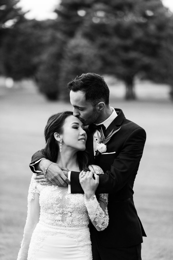 Black-and-white photo of the groom embracing the bride from behind on the lawn.
