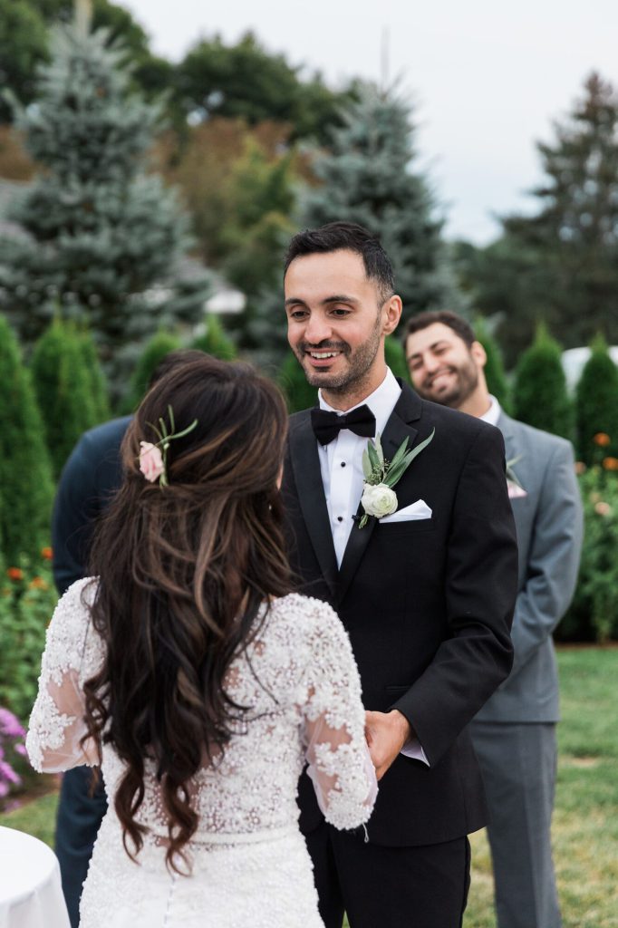 Groom smiling at the bride during the ceremony as his best man looks on.