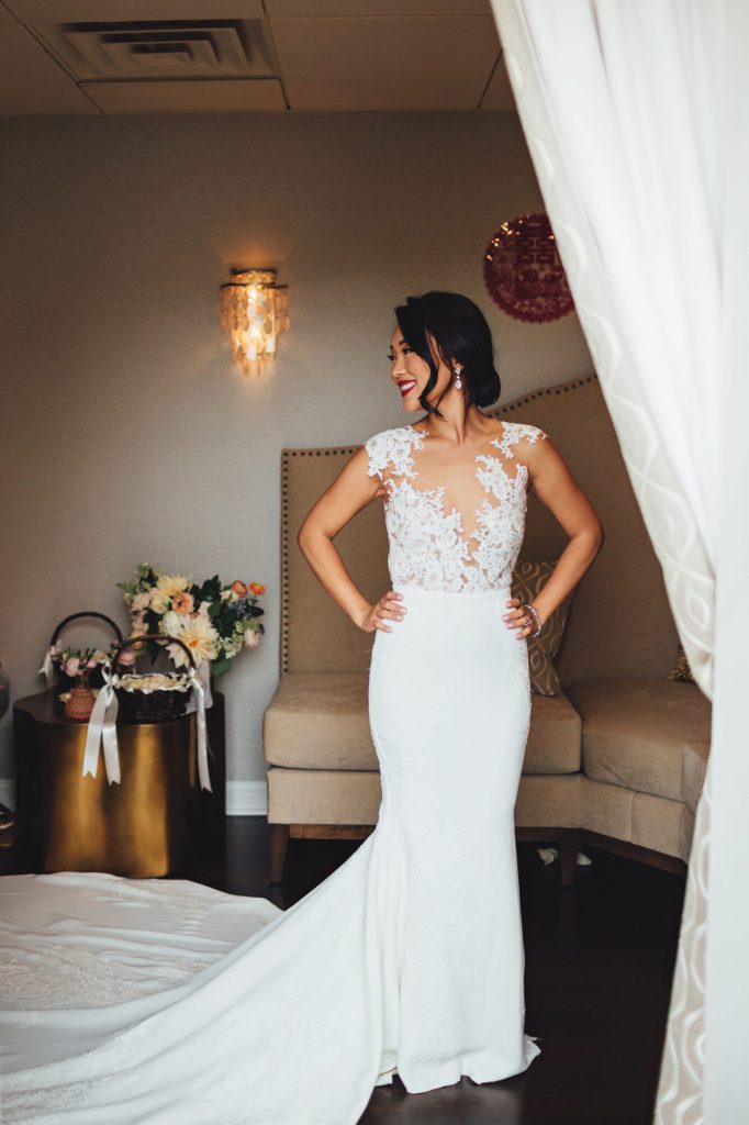 Bride in a fitted lace gown posing in a lounge doorway framed by white drapes.