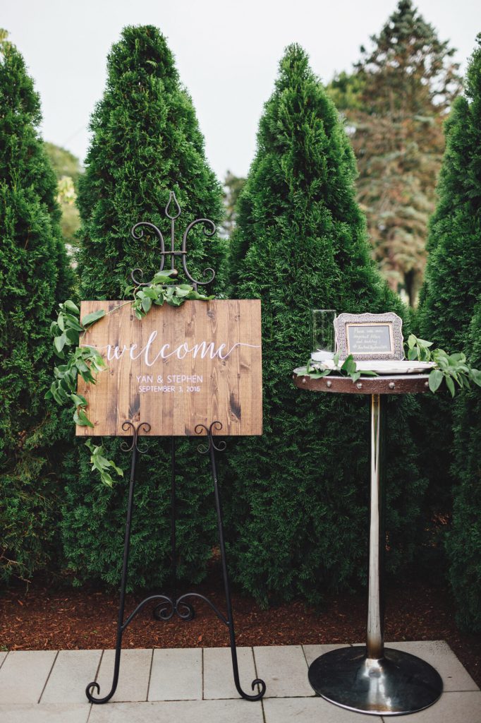 Wooden welcome sign on an easel beside a card table in front of tall evergreens.