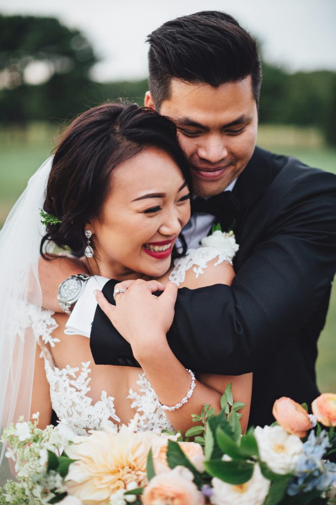 Groom hugging the laughing bride from behind as she holds her bouquet.