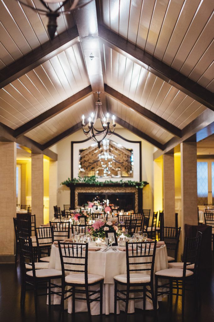 Reception hall with a vaulted beamed ceiling and chandelier over tables with pink centerpieces.