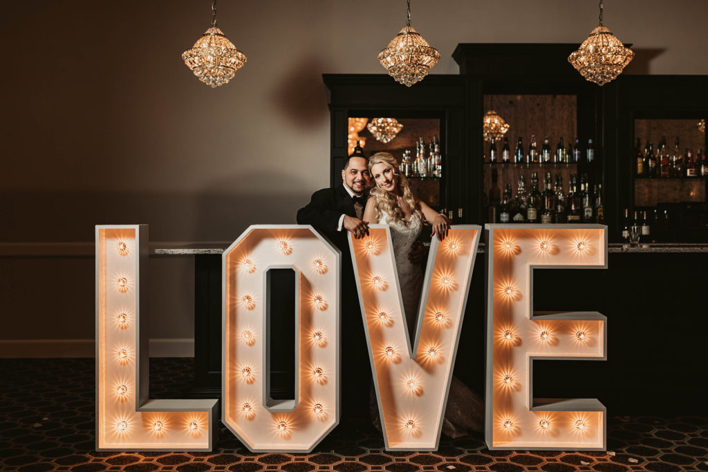 Marquee Letters Spelling Love near Bar with Bride and Groom Smiling