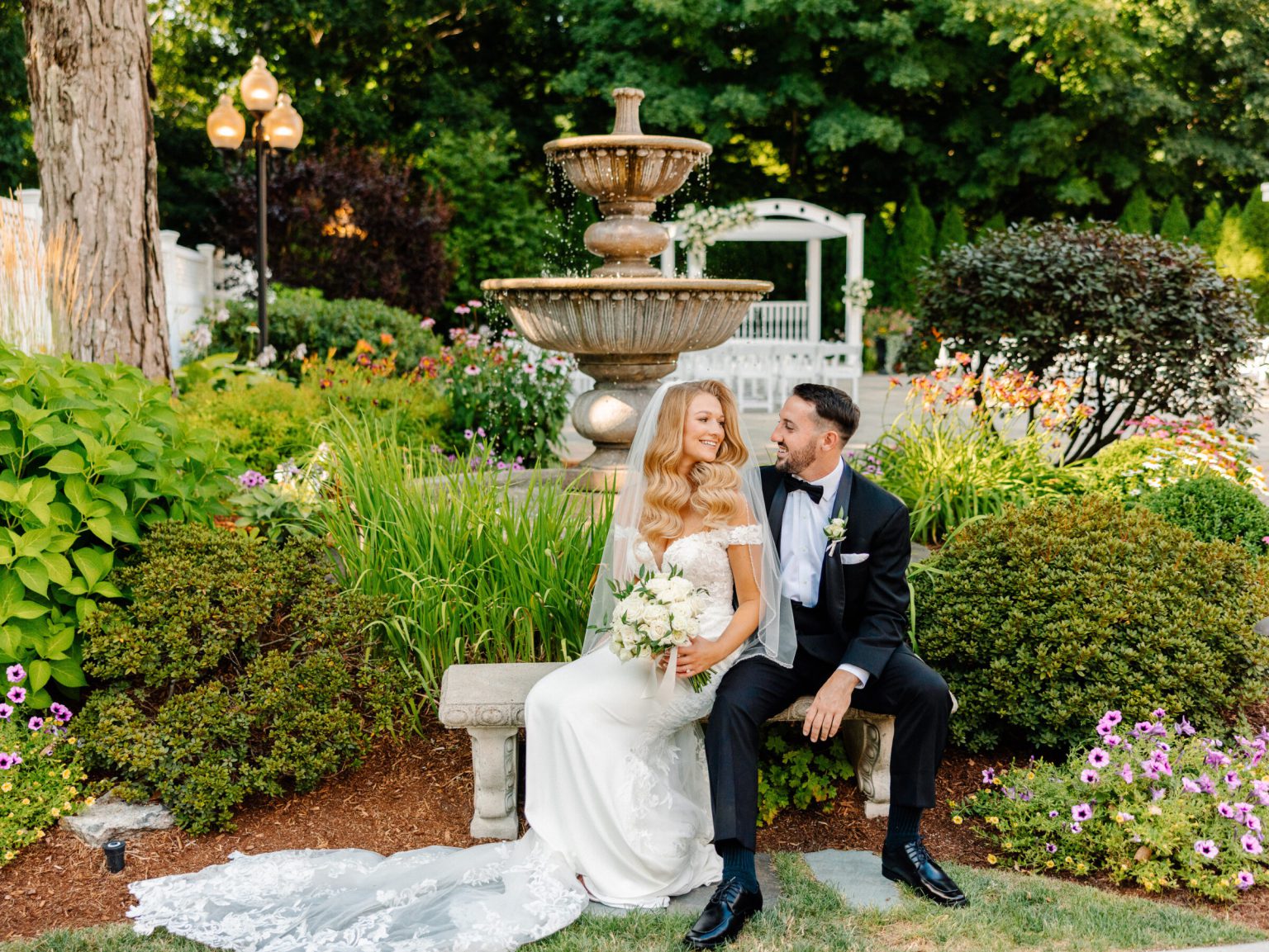 Bride in an off-the-shoulder gown and groom in a tuxedo seated on a stone bench in a garden, before a tiered fountain, lamp posts and flower beds.