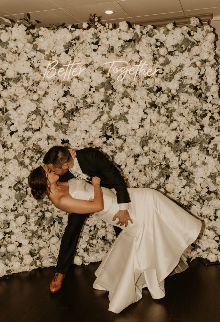 Bride and groom kiss in front of flower wall and neon sign at The Villa in East Bridgewater, Massachusetts