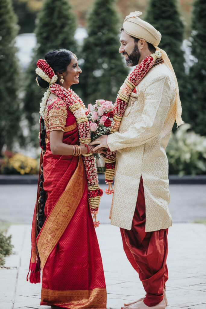 Bride and Groom Wearing a Red Sari and Beige Sherwani Hold Hands Outside at Avenir