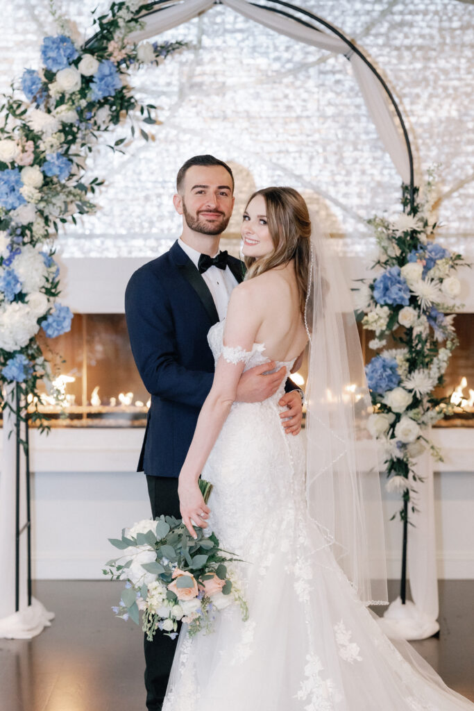 Bride and Groom in Front of Fireplace Under Dusty Blue and White Floral Arch