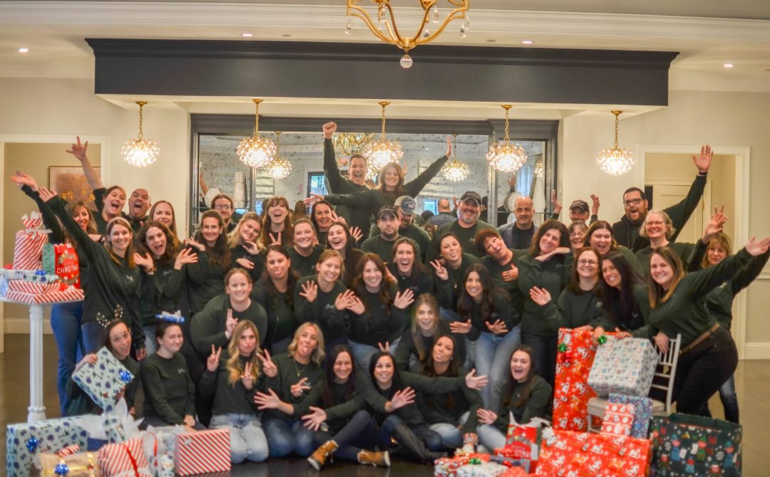 Large group of volunteers cheering together behind piles of wrapped gifts.