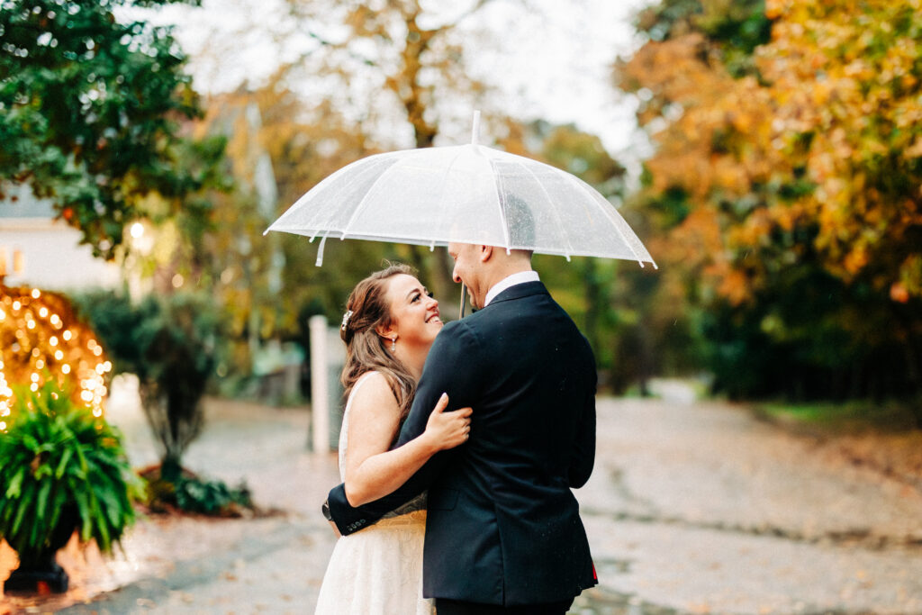 Bride and Groom Under Umbrella in Front of Saphire Estate Fall Foliage