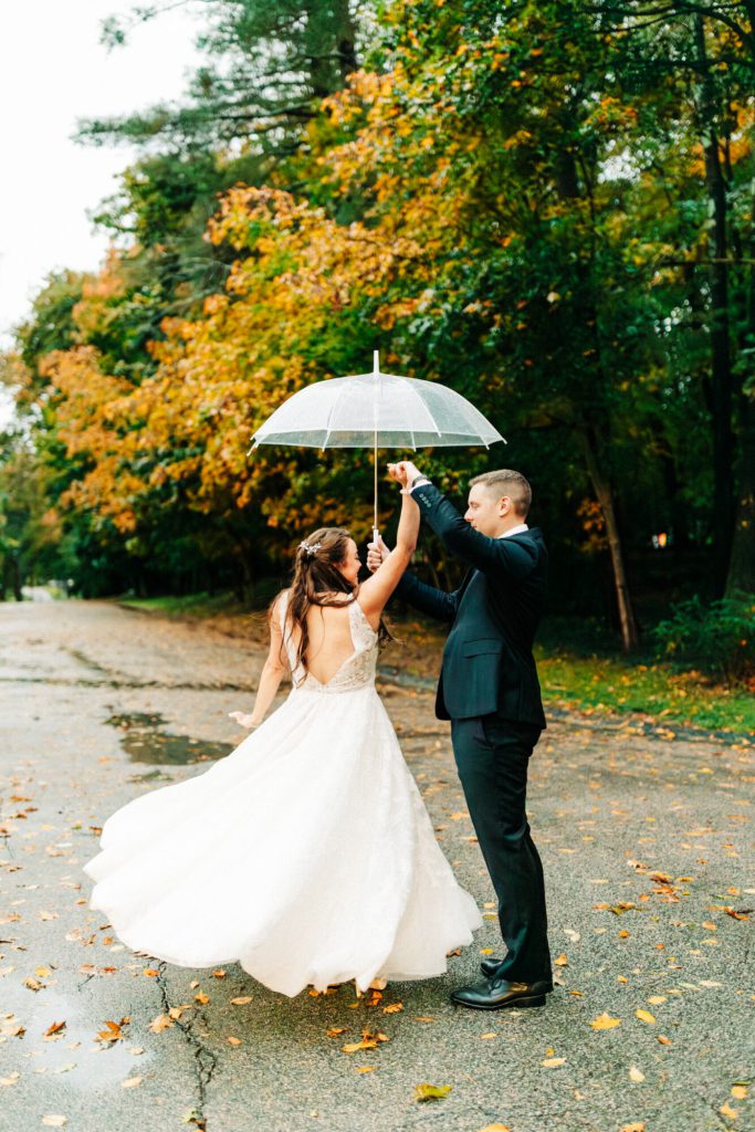Groom Twirls Bride Under Umbrella With Fall Foliage Backdrop