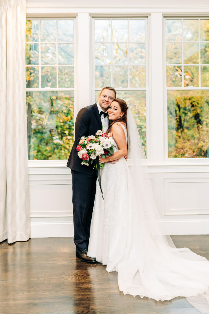 Bride and Groom Pose in Front of Fall Foliage Behind Window