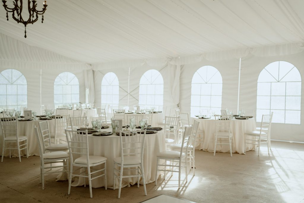 White tent with arched windows, chandeliers and round tables set for dinner.