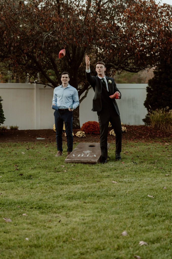 Groom and a guest playing cornhole on a lawn.