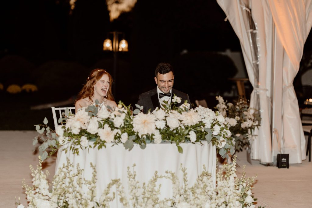 Bride and groom at a sweetheart table with white florals in a tent.