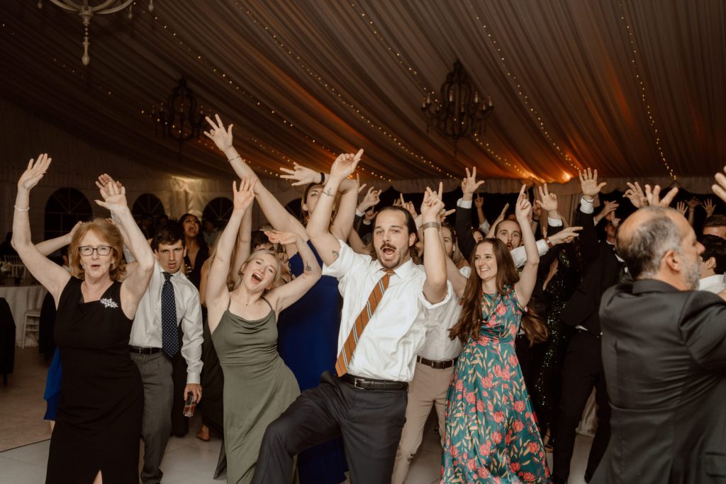Guests dancing with arms raised under chandeliers in a tent.