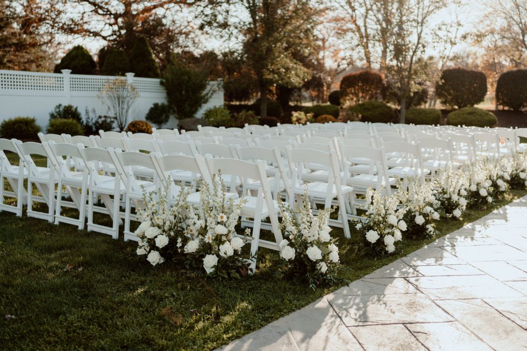 White ceremony chairs with white florals lining a stone aisle.