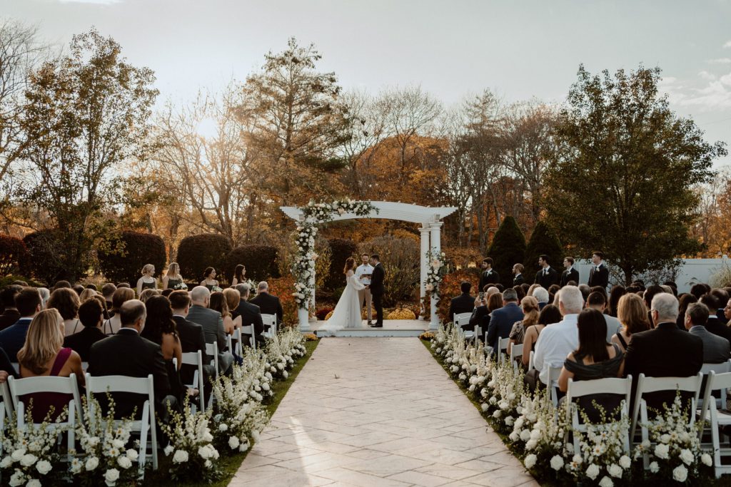 Guests watching a ceremony beneath a white pergola in a fall garden.