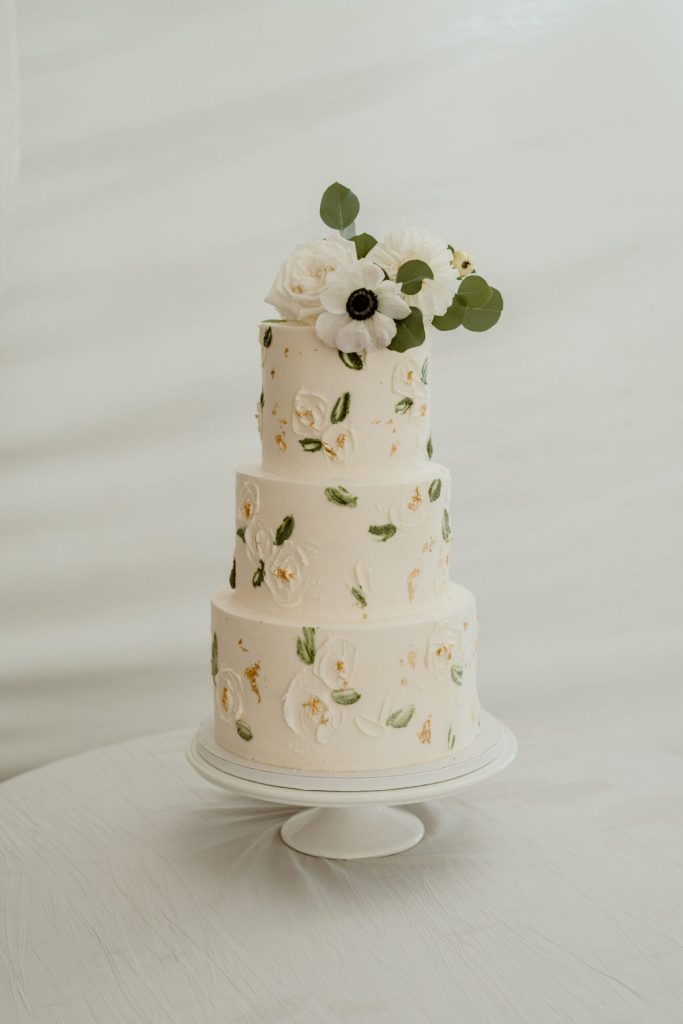 Three-tier white wedding cake with pressed greenery and white flowers.
