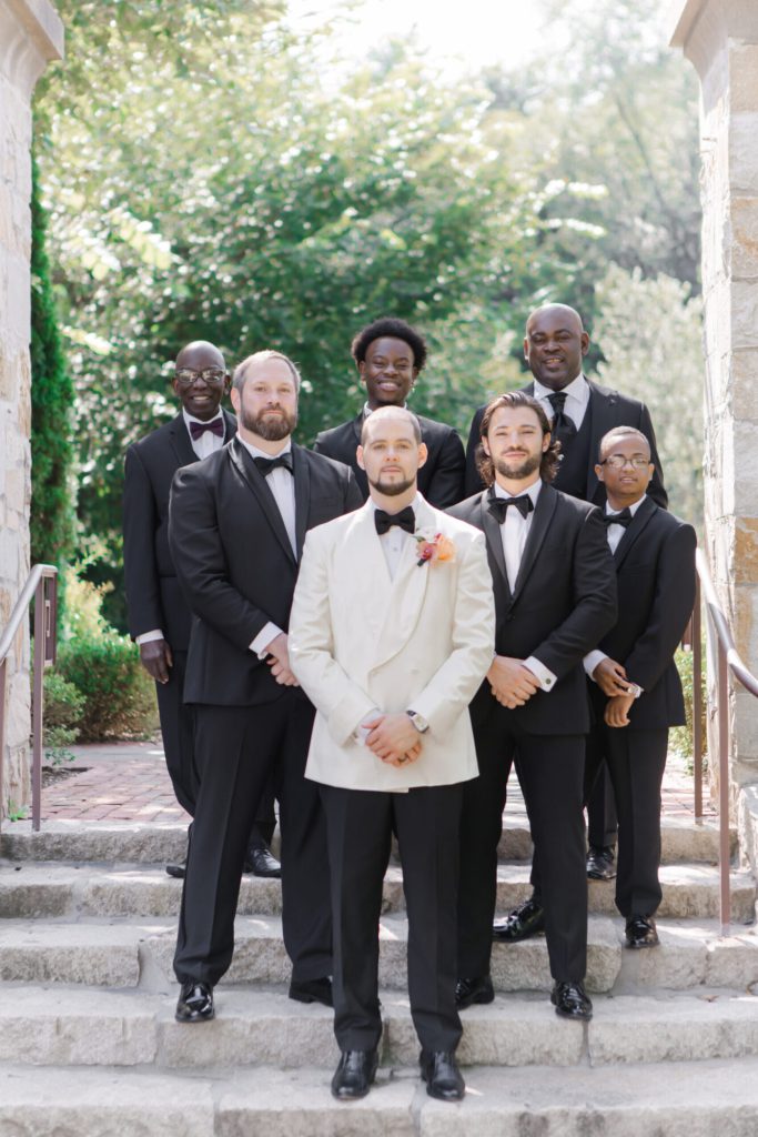 Groom in a white jacket with groomsmen in black on stone steps.