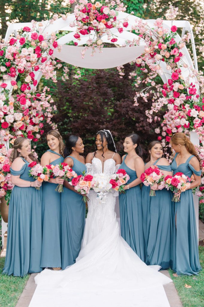 Bride with bridesmaids in teal gowns before a pink floral arch.