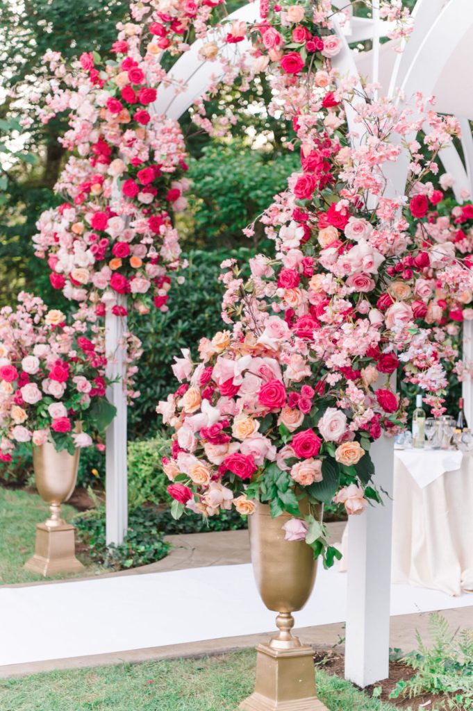 Lush pink and white floral arch on gold stands in a garden.