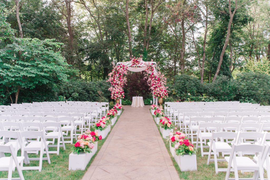 Garden ceremony with white chairs facing a pink floral arch among trees.