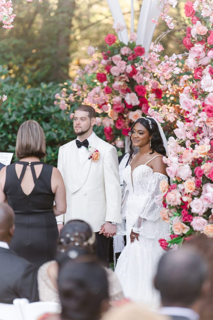 Bride and groom beneath a pink floral arch with the officiant.