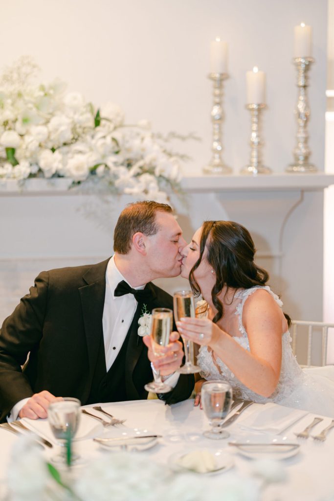 Bride and groom kissing while toasting by a candlelit mantel.