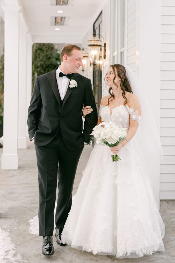 Bride and groom walking arm in arm along a white porch.