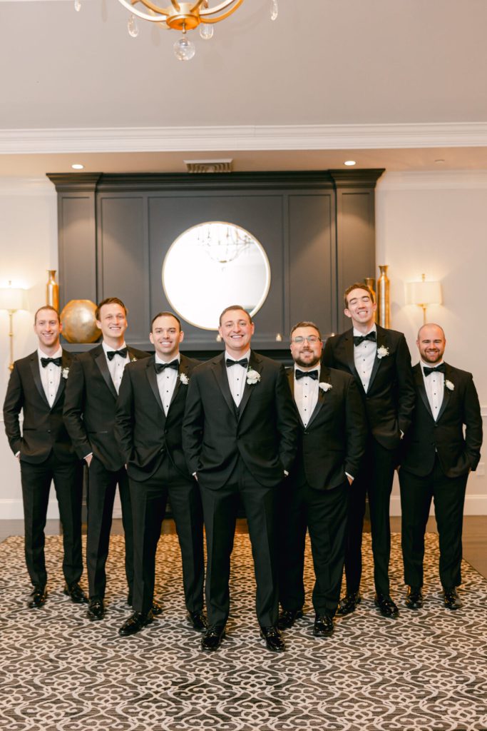 Groom with groomsmen in black tuxedos before a dark wall.
