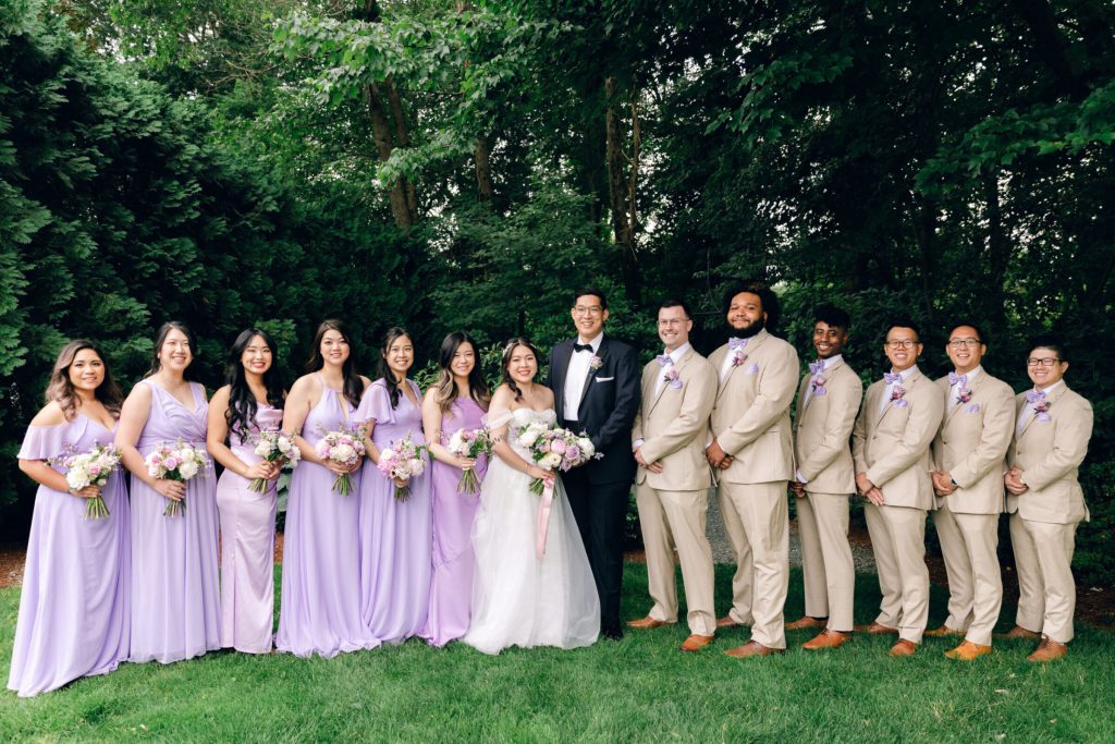 Wedding party in lavender gowns and tan suits lined up on a lawn.