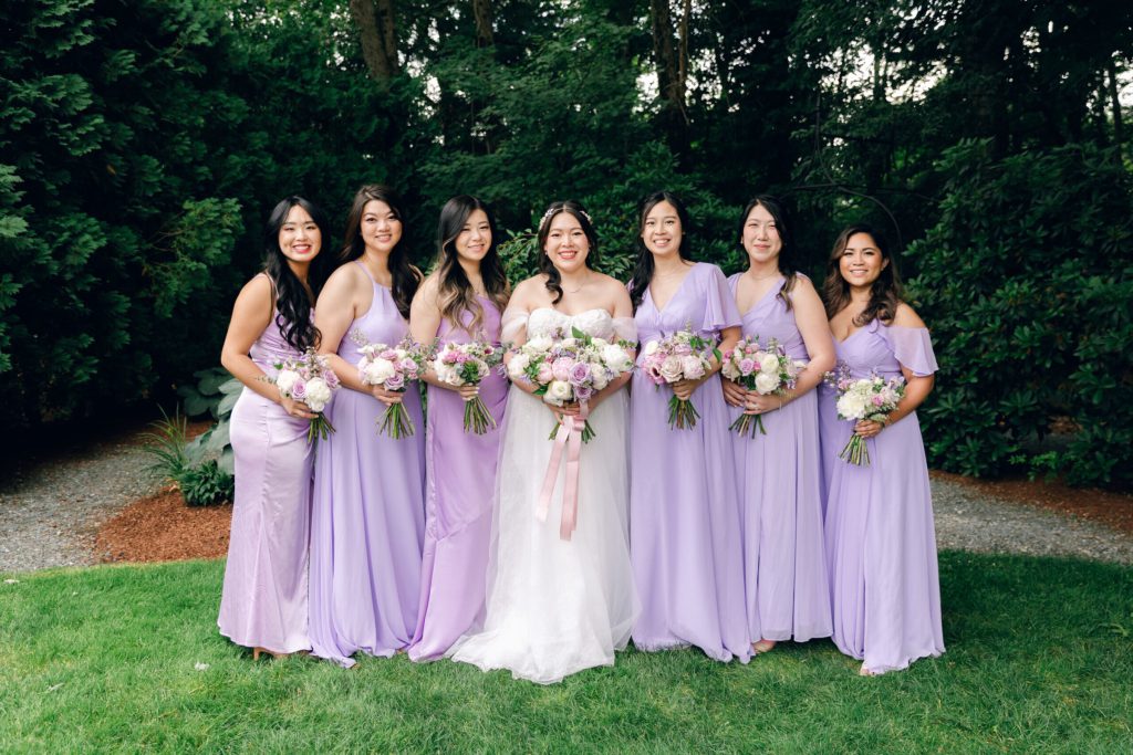 Bride with bridesmaids in lavender gowns on a lawn.
