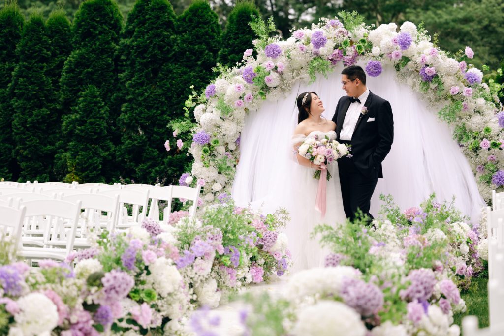 Bride and groom beneath a lavender floral arch on a lawn.