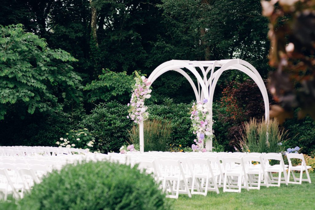 White garden arch with florals and rows of white chairs.