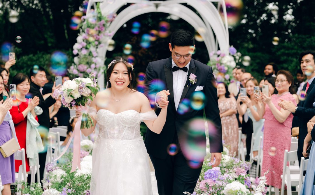 Bride and groom walking back up the aisle through a shower of bubbles under a white arch draped in purple and white flowers as guests cheer.