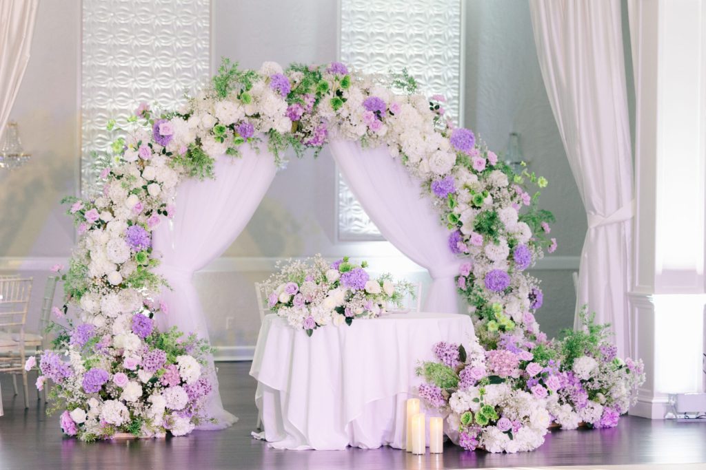 Lavender and white floral arch over a sweetheart table in a draped ballroom.