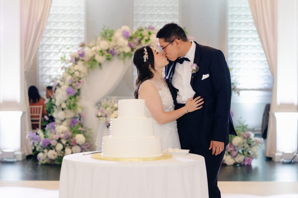Bride and groom kissing beside their wedding cake under a floral arch.