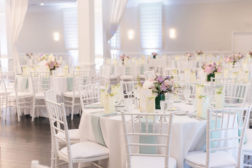 White ballroom with round tables and lavender centerpieces.
