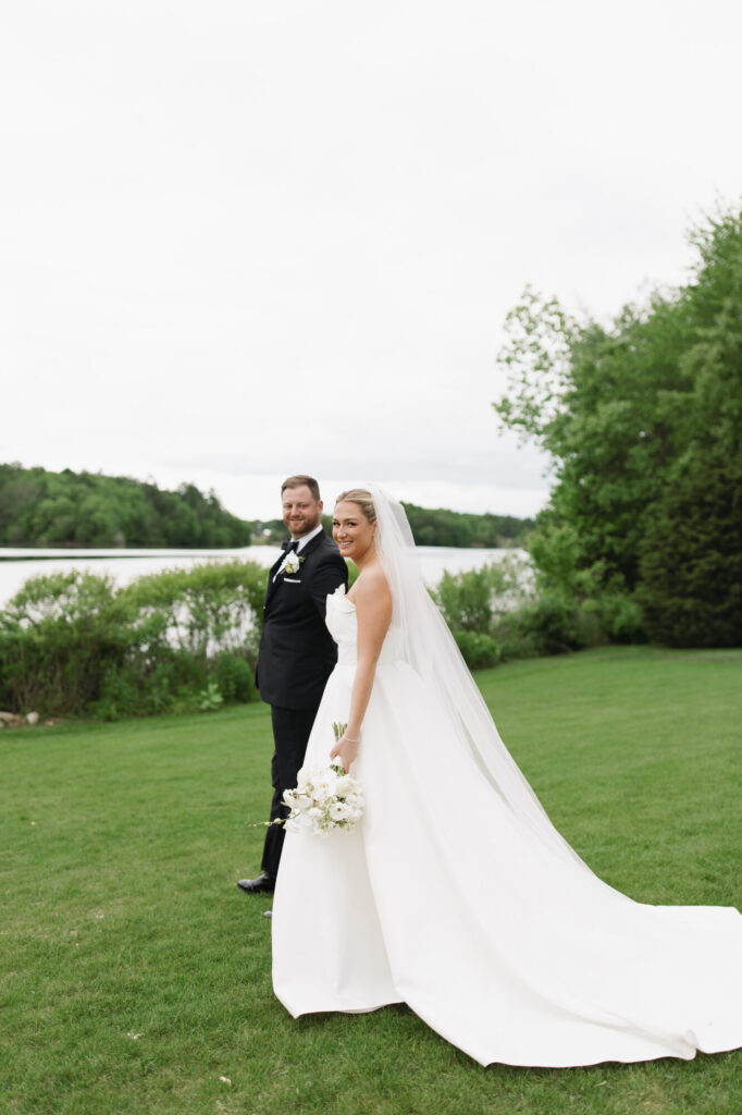 Bride and groom pose by lake and lush greenery backdrop