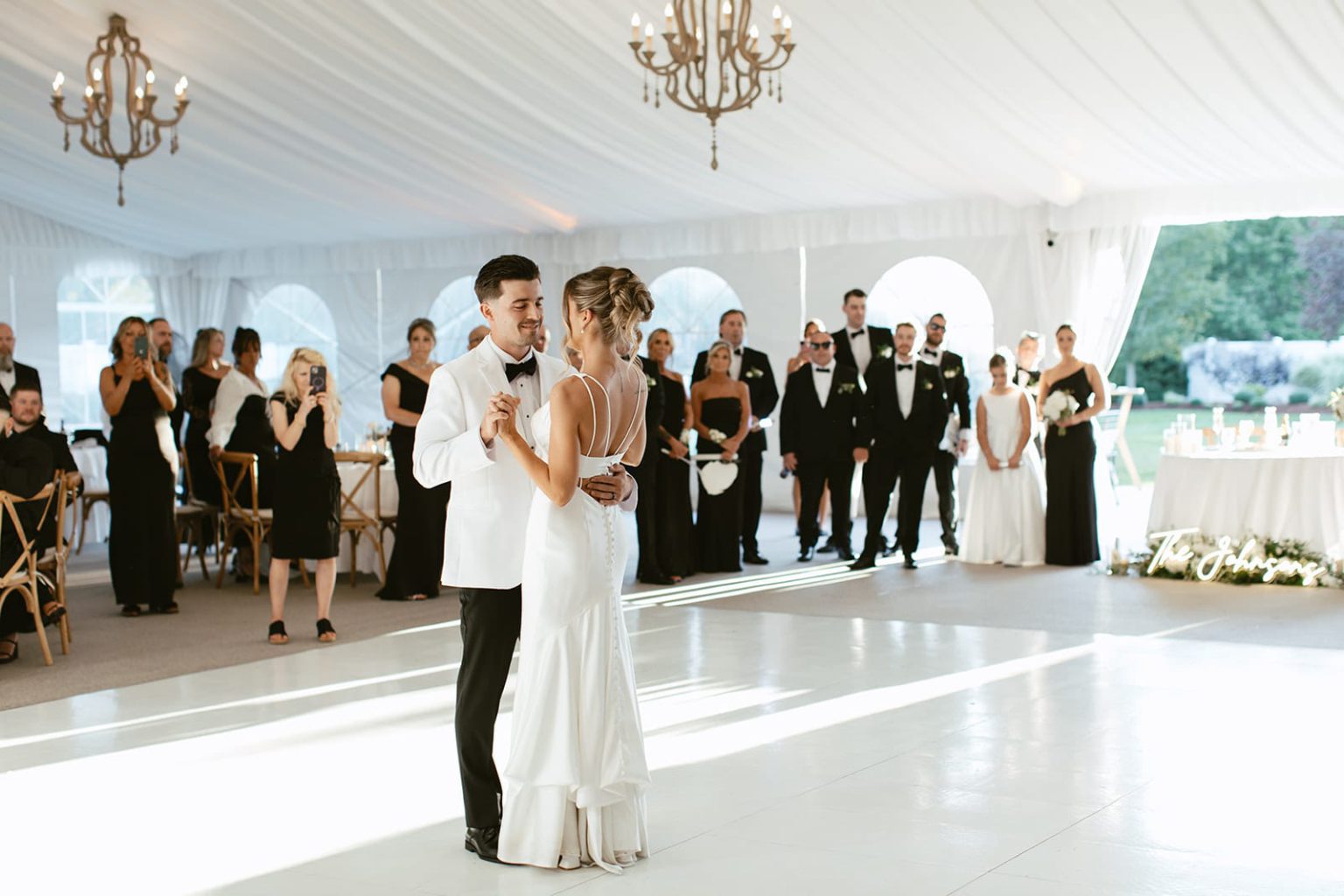 First dance on a white floor in The Villa's tent under brass chandeliers, the bride in a satin gown and groom in a white jacket, as guests watch.