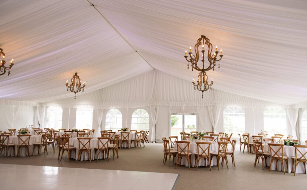 White draped reception tent with wrought iron candle chandeliers, round tables in white linens with wooden cross-back chairs and arched windows.