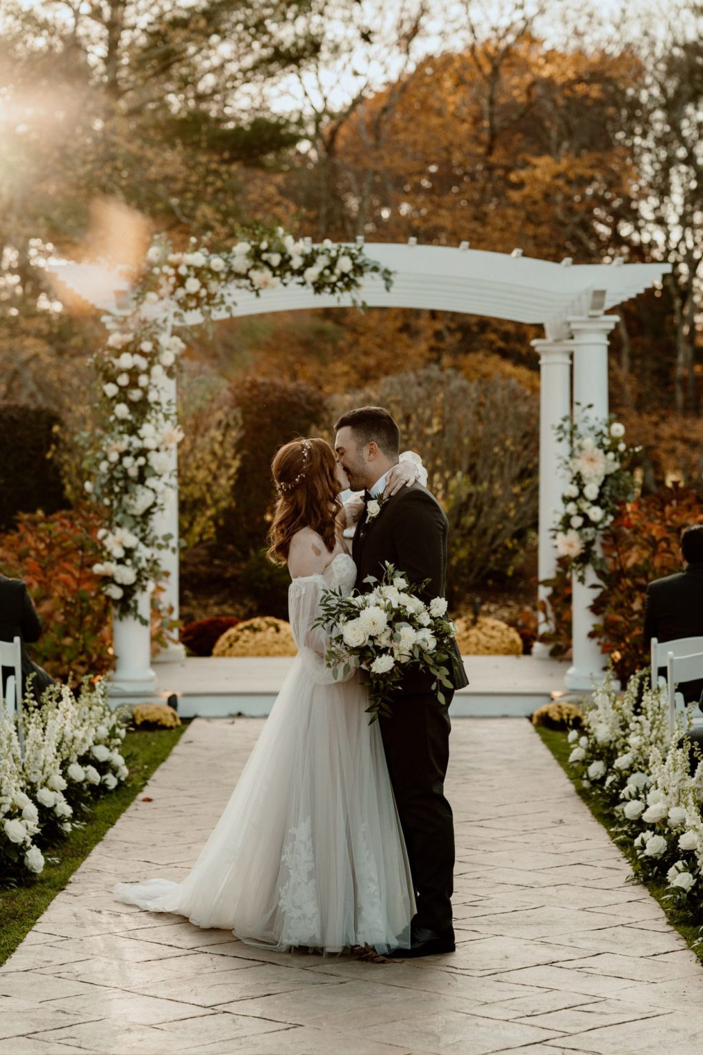 Couple kissing beneath a flower-covered white pergola at an autumn ceremony