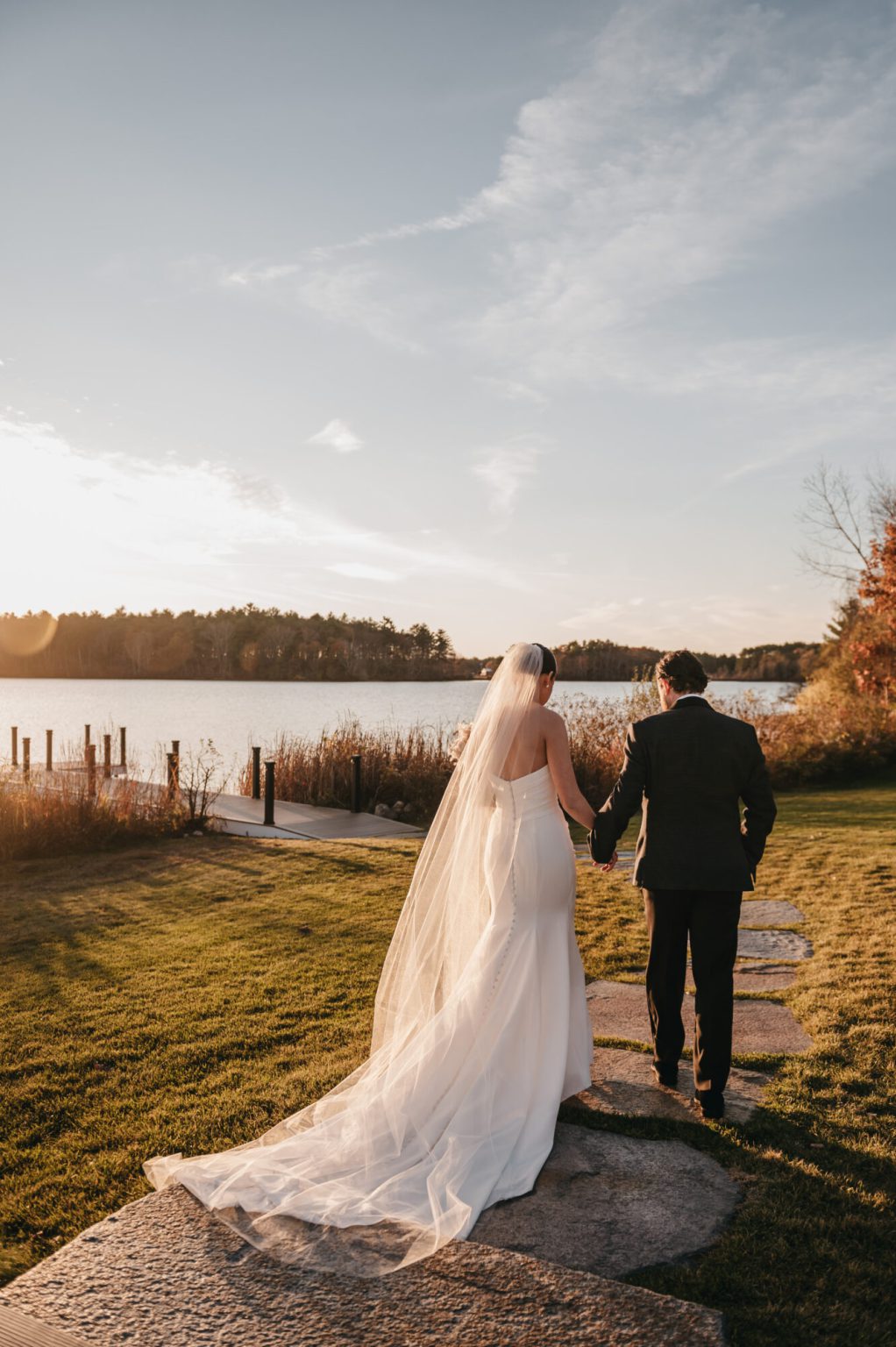 Bride and groom walking hand in hand toward the lake at sunset