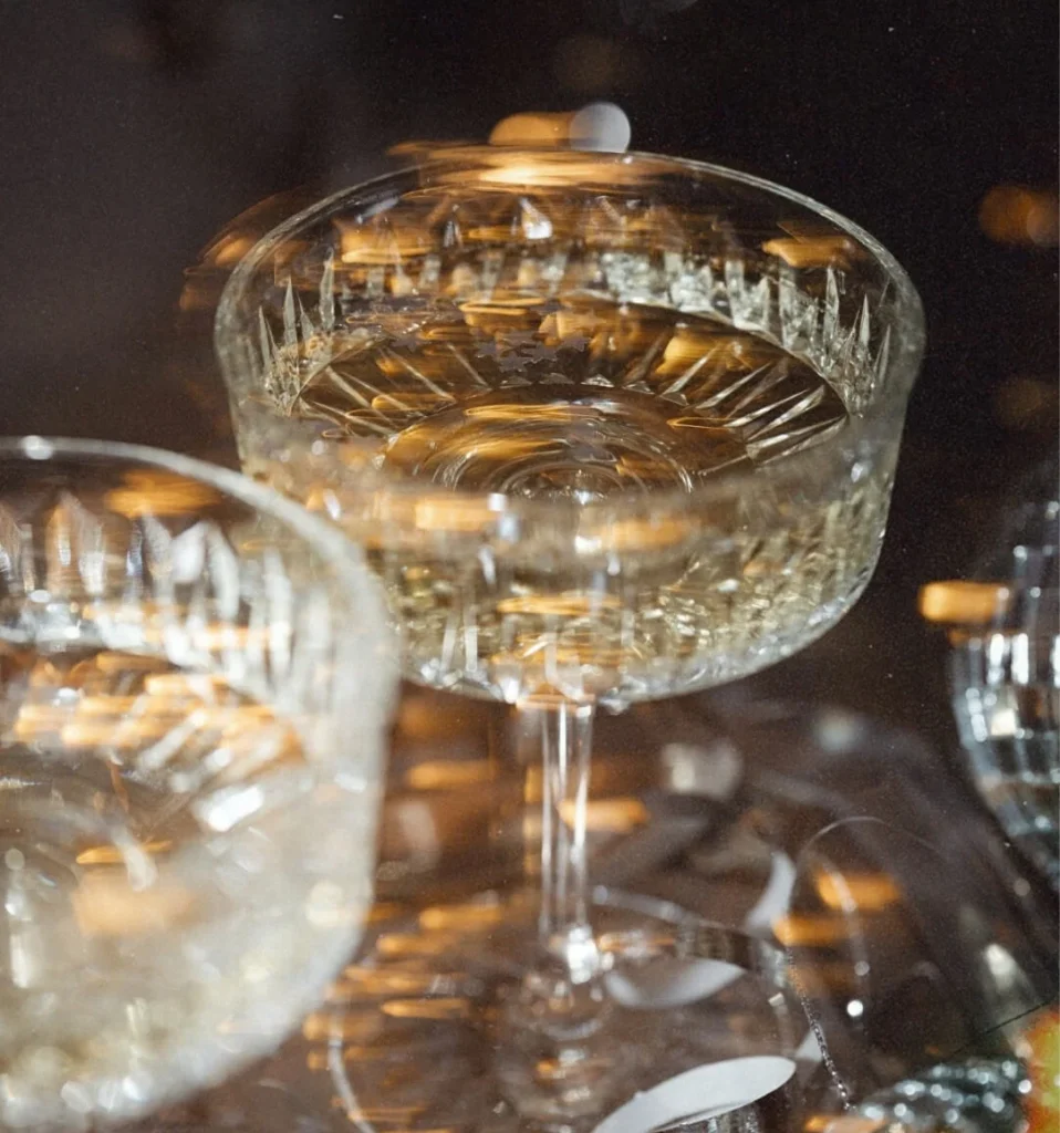 Close-up of a crystal champagne coupe filled with sparkling wine beside a second glass, with warm bokeh lights glowing on a dark background.
