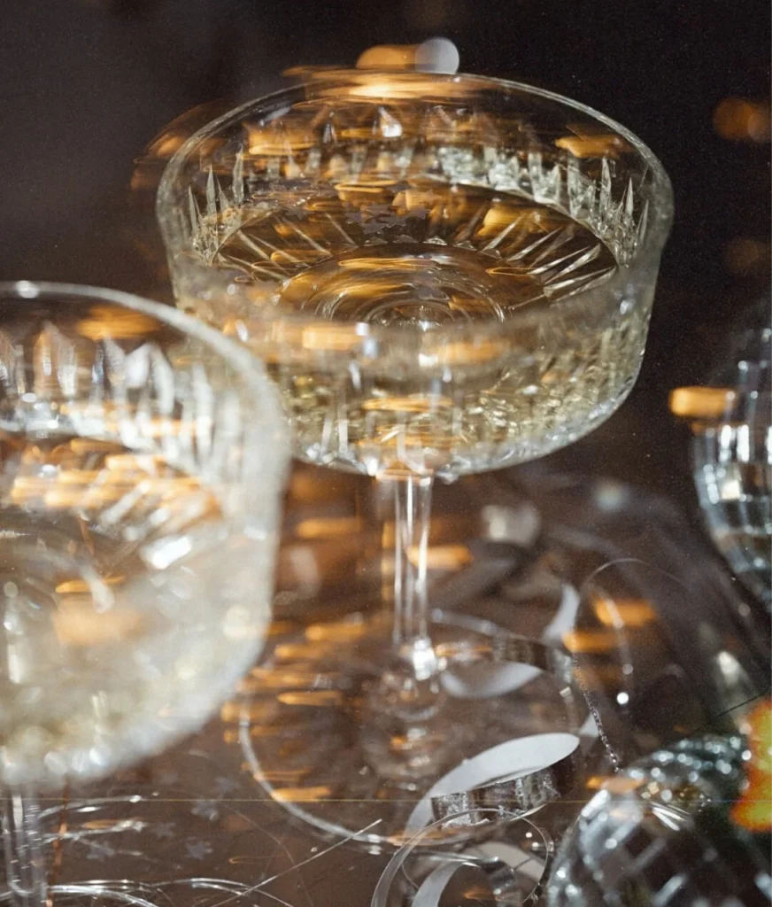 Close-up of a crystal champagne coupe filled with sparkling wine beside a second glass, with warm bokeh lights glowing on a dark background.