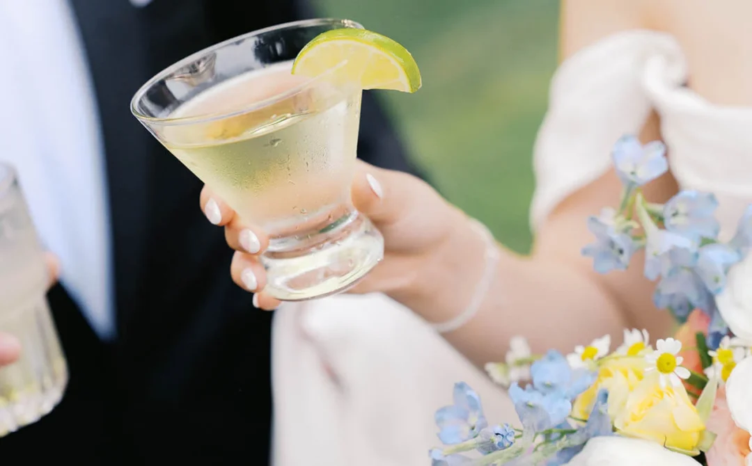 Bride in an off-shoulder gown holding a stemless martini glass with a lime wedge and a bouquet of blue, yellow and white flowers by the groom.